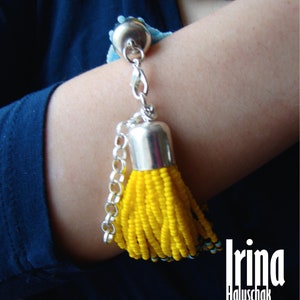 May include: A yellow beaded bracelet with a silver tassel charm and a silver chain. The bracelet is worn on a person's wrist. The text "Irina Haluschak" is visible in the image.