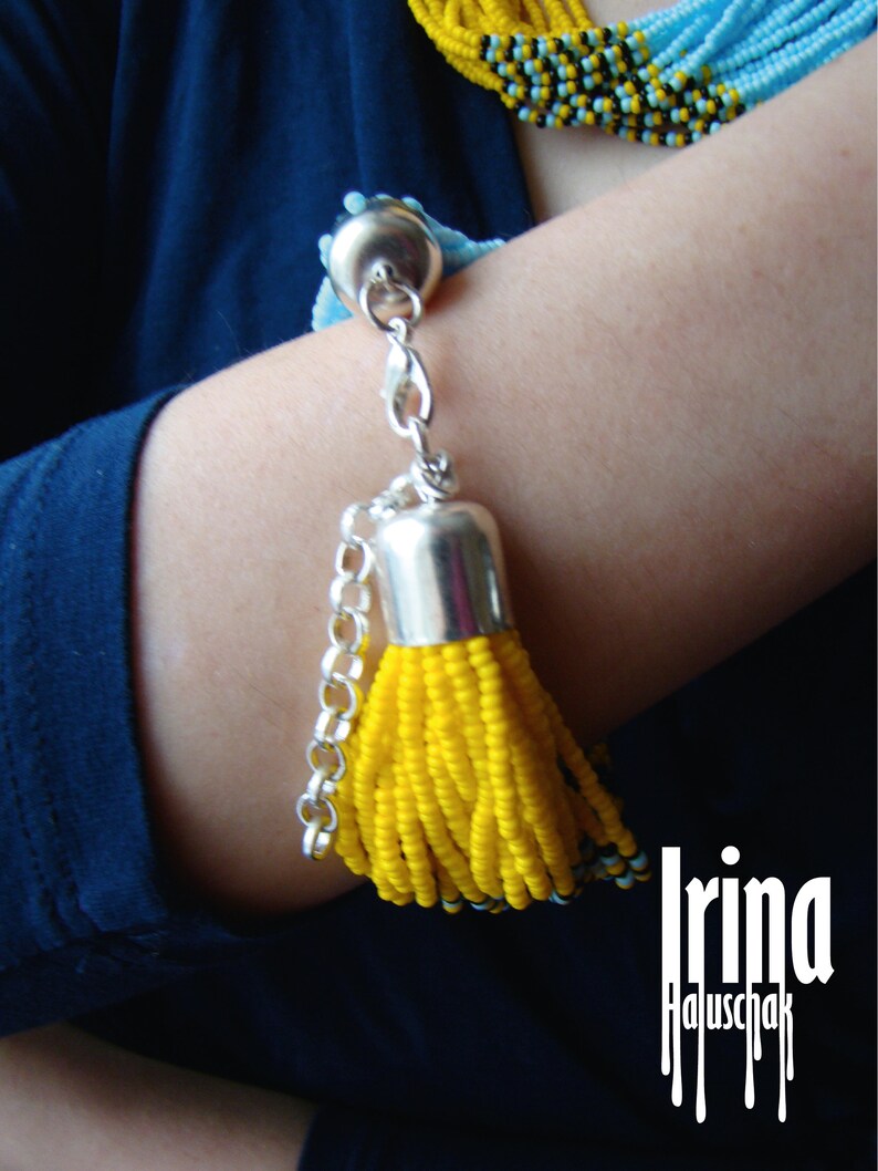 May include: A yellow beaded bracelet with a silver tassel charm and a silver chain. The bracelet is worn on a person's wrist. The text "Irina Haluschak" is visible in the image.