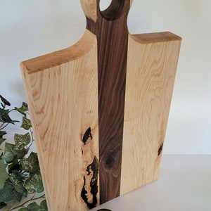 Könnte beinhalten: Ein großes Schneidebrett aus Holz mit einem Griff. Das Brett ist aus hellem Holz mit einem dunklen Holzstreifen in der Mitte. In der rechten unteren Ecke des Bildes befinden sich drei Limetten.