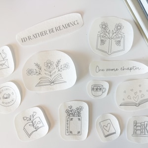 Peut inclure: Un ensemble de onze autocollants blancs avec des illustrations au trait noir représentant des livres, des fleurs, des cœurs et une tasse de café. Le texte inclut I'D RATHER BE READING, One more chapter et HOT GIRLS READ BOOKS. Formes irrégulières.