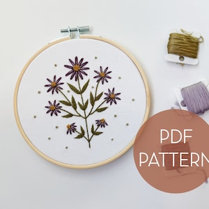 Peut inclure: Un cerceau à broder avec un motif floral composé de fleurs violettes et de feuilles vertes. Le cerceau est sur un fond blanc avec deux bobines de fil à broder, une verte et une violette, en arrière-plan. Le texte "PDF PATTERN" est écrit dans un cercle sur le côté droit de l'image.