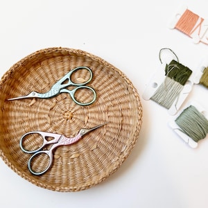 Puede incluir: Dos pares de tijeras de bordado en forma de pájaro en una cesta tejida. Las tijeras son de diferentes colores, una es turquesa y la otra rosa. También hay carretes de hilo en varios tonos de verde y rosa.