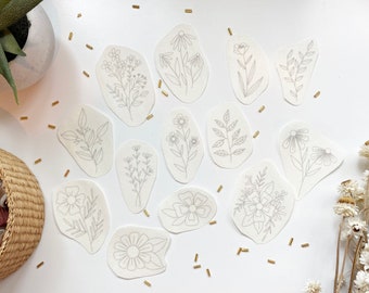 May include: A set of 12 white paper cutouts with various floral designs. The cutouts are in the shape of irregular ovals and feature simple line drawings of flowers and leaves.
