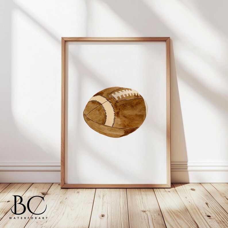 May include: A watercolor painting of a brown football with white stitching. The football is centered on a white background and framed in a light brown frame.