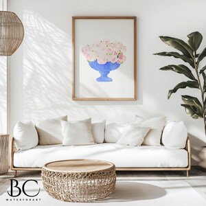 May include: A watercolour painting of a blue vase filled with pink flowers. The painting is framed and hanging on a white wall above a white sofa with cushions. A woven coffee table sits in front of the sofa.