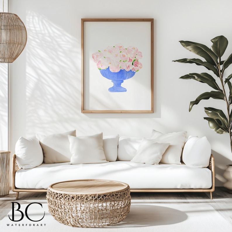 May include: A watercolour painting of a blue vase filled with pink flowers. The painting is framed and hanging on a white wall above a white sofa with cushions. A woven coffee table sits in front of the sofa.