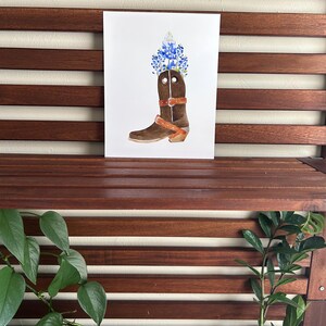 Puede incluir: Una pintura de acuarela de una bota vaquera marr&oacute;n con flores azules y blancas en la parte superior. La bota tiene una correa de cuero marr&oacute;n en la parte superior.
