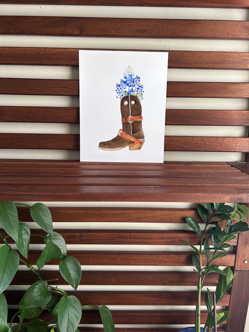 Puede incluir: Una pintura de acuarela de una bota vaquera marr&oacute;n con flores azules y blancas en la parte superior. La bota tiene una correa de cuero marr&oacute;n en la parte superior.