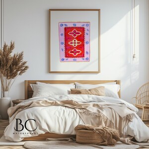 May include: A watercolor painting of a pink and red rug with a floral design. The painting is framed and hanging on a white wall above a bed with white bedding and a tan throw blanket.