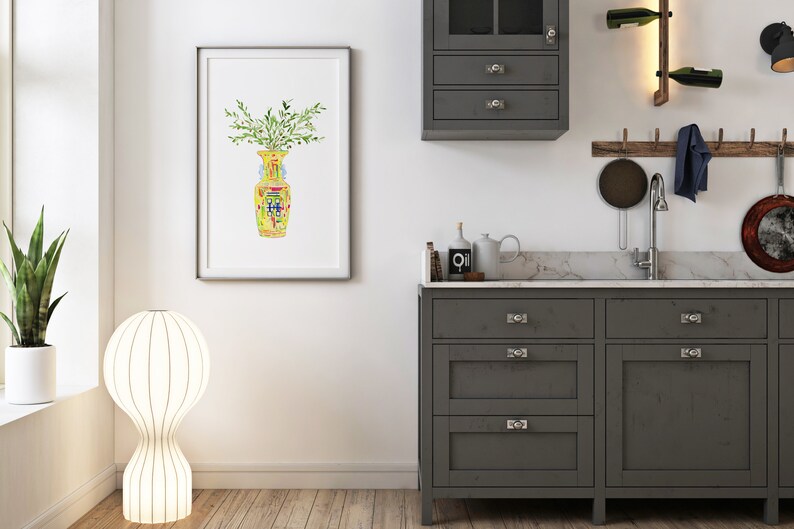 May include: A watercolor painting of a yellow and green vase with a floral design. The vase is filled with green leaves and is framed in a white frame.