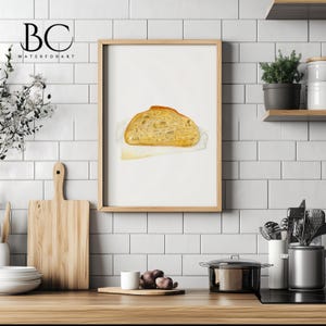 May include: A watercolor painting of a loaf of bread on a white napkin. The bread is light brown and has a slightly crusty exterior. The painting is framed in a light wood frame and is hanging on a white tiled wall.