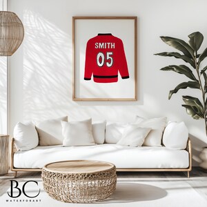 Puede incluir: Una ilustraci&oacute;n enmarcada de una camiseta roja con ribetes negros y el nombre "SMITH" y el n&uacute;mero "05" en la espalda. La camiseta est&aacute; colgada en una pared blanca sobre un sof&aacute; blanco.