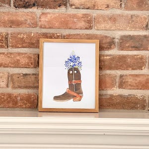 Puede incluir: Una pintura de acuarela enmarcada de una bota vaquera marr&oacute;n con flores azules en la parte superior. La bota tiene una correa marr&oacute;n y una hebilla plateada.