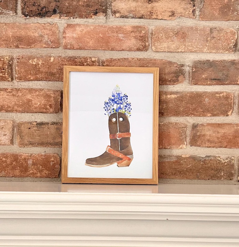 Puede incluir: Una pintura de acuarela enmarcada de una bota vaquera marr&oacute;n con flores azules en la parte superior. La bota tiene una correa marr&oacute;n y una hebilla plateada.