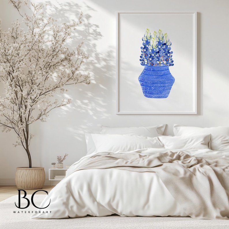 May include: A watercolor painting of a blue vase with a geometric pattern filled with blue flowers. The painting is framed and hanging on a white wall above a white bed with a beige blanket.
