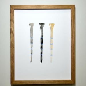 May include: Three watercolor golf tees in a wooden frame. The tees are gray, black, and beige with blue stripes.