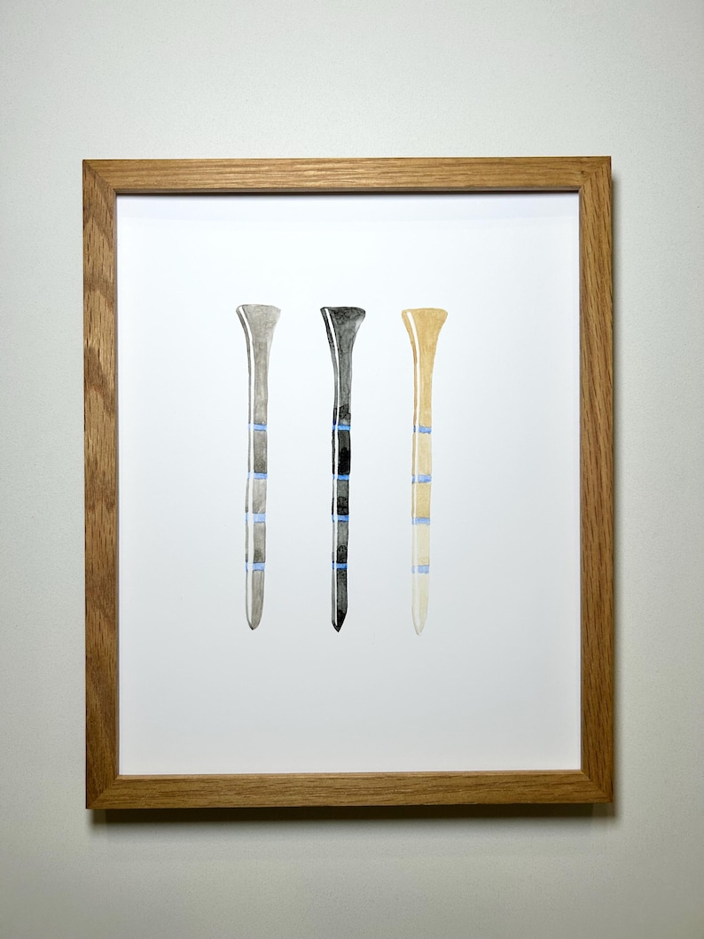 May include: Three watercolor golf tees in a wooden frame. The tees are gray, black, and beige with blue stripes.