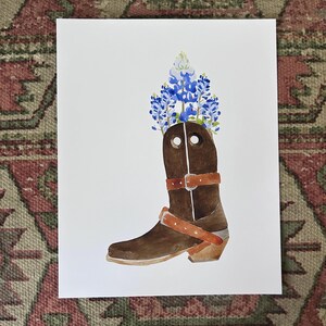 Puede incluir: Una pintura de acuarela de una bota vaquera marr&oacute;n con un ramo de flores azules y blancas de lupino de Texas en la parte superior de la bota.