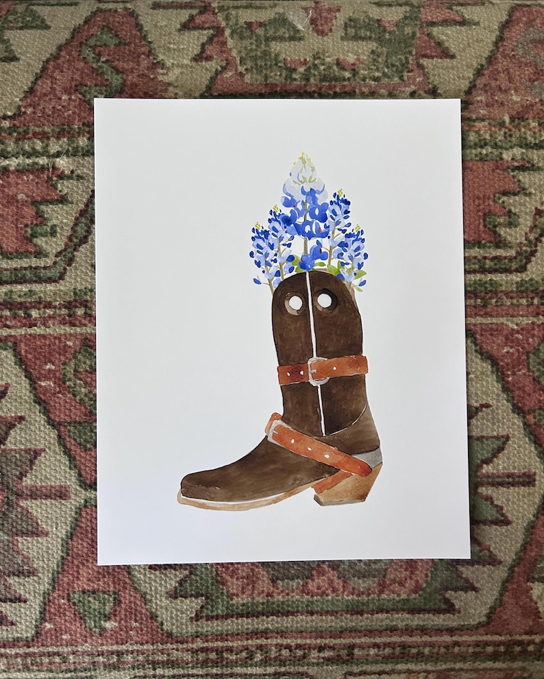 Puede incluir: Una pintura de acuarela de una bota vaquera marr&oacute;n con un ramo de flores azules y blancas de lupino de Texas en la parte superior de la bota.