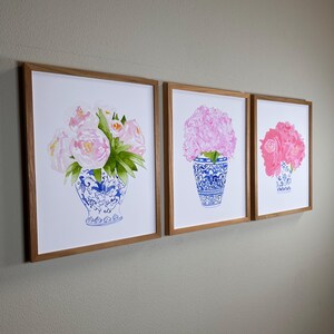 Puede incluir: Tres impresiones enmarcadas de flores rosas en jarrones azules y blancos. Los jarrones tienen un dise&ntilde;o cl&aacute;sico azul y blanco, y los marcos son de madera clara. Las impresiones se exhiben en una pared.