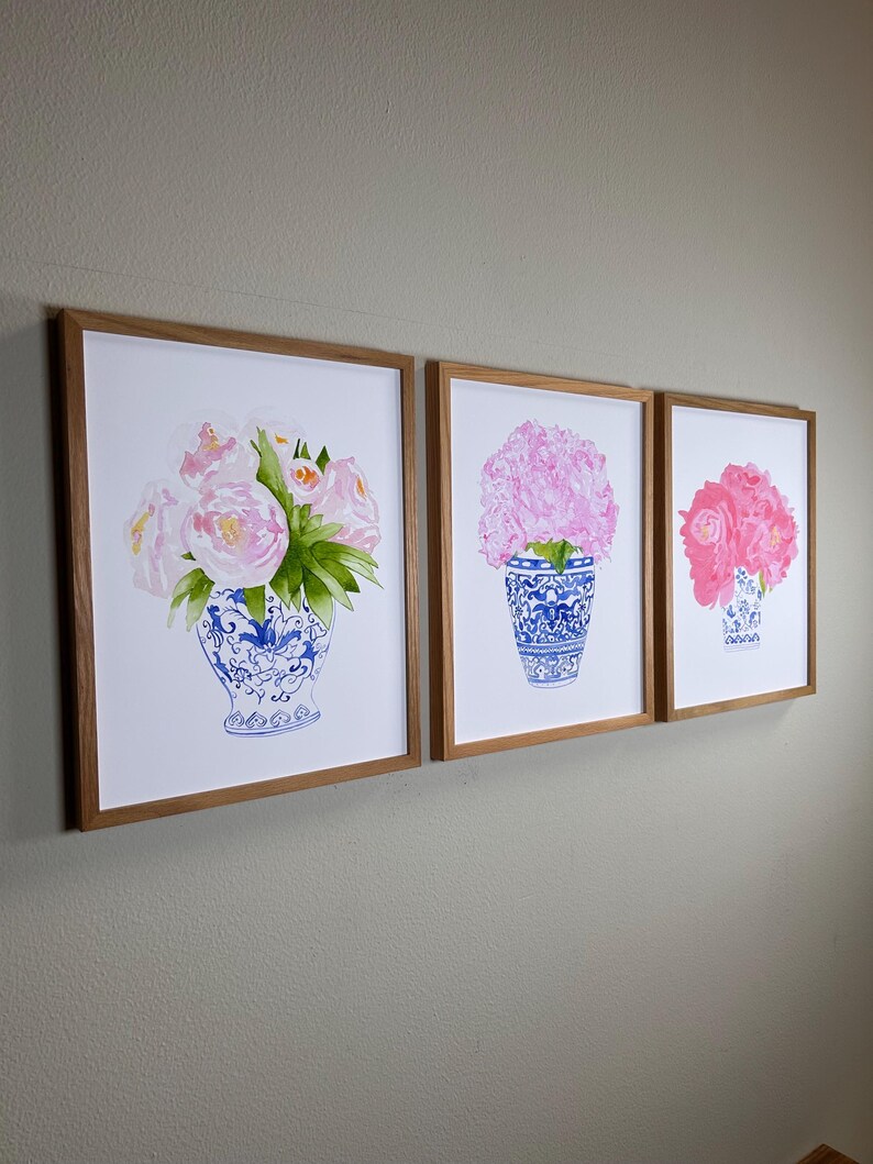 Puede incluir: Tres impresiones enmarcadas de flores rosas en jarrones azules y blancos. Los jarrones tienen un dise&ntilde;o cl&aacute;sico azul y blanco, y los marcos son de madera clara. Las impresiones se exhiben en una pared.