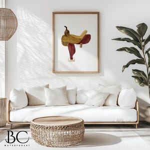 May include: A watercolor painting of a saddle in shades of brown and red, framed in a natural wood frame. The painting is hanging on a white wall above a white couch with beige throw pillows.