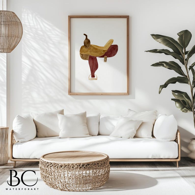 May include: A watercolor painting of a saddle in shades of brown and red, framed in a natural wood frame. The painting is hanging on a white wall above a white couch with beige throw pillows.