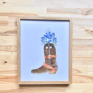 Puede incluir: Una pintura de acuarela de una bota vaquera marr&oacute;n con flores azules y blancas en la parte superior. La bota tiene una correa y una hebilla marrones.