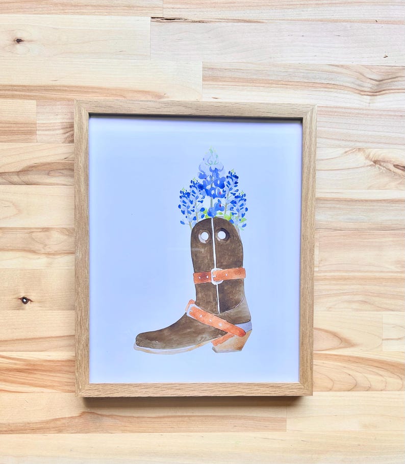 Puede incluir: Una pintura de acuarela de una bota vaquera marr&oacute;n con flores azules y blancas en la parte superior. La bota tiene una correa y una hebilla marrones.