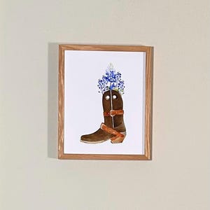 Puede incluir: Una pintura de acuarela de una bota vaquera marr&oacute;n con flores azules en la parte superior. La bota tiene una correa de cuero marr&oacute;n y una hebilla.