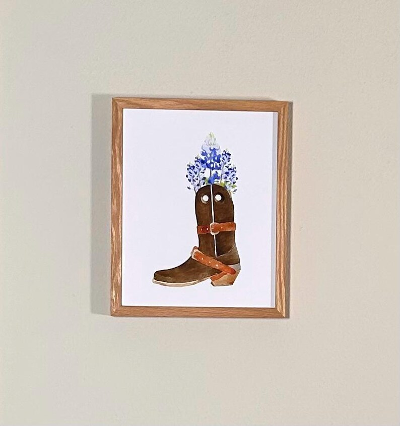 Puede incluir: Una pintura de acuarela de una bota vaquera marr&oacute;n con flores azules en la parte superior. La bota tiene una correa de cuero marr&oacute;n y una hebilla.