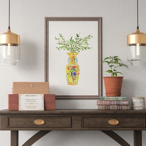 May include: A framed watercolor painting of a yellow vase with green leaves and red, blue, and yellow accents. The vase is filled with green leaves and is sitting on a table with two gold pendant lights hanging above it.