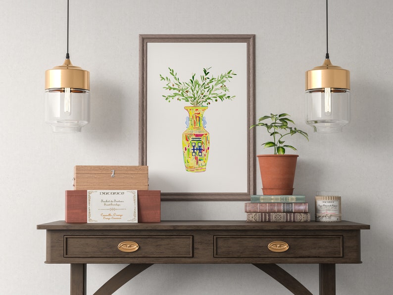May include: A framed watercolor painting of a yellow vase with green leaves and red, blue, and yellow accents. The vase is filled with green leaves and is sitting on a table with two gold pendant lights hanging above it.