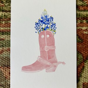 Puede incluir: Una bota vaquera rosa con espuela, decorada con flores de campanilla azul y blanca de Texas.
