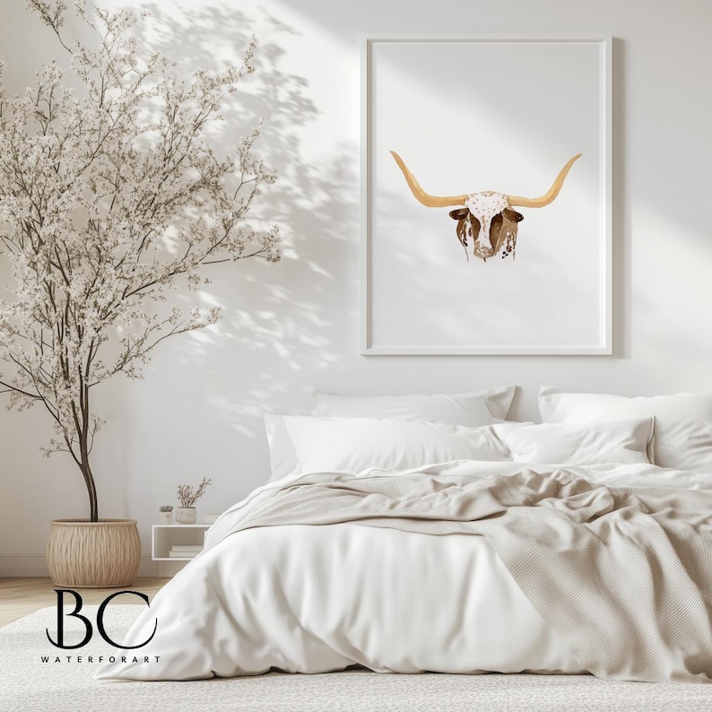 May include: A white framed print of a watercolor painting of a longhorn steer head with brown horns and white spots. The print is hanging on a white wall above a white bed with a tan blanket.