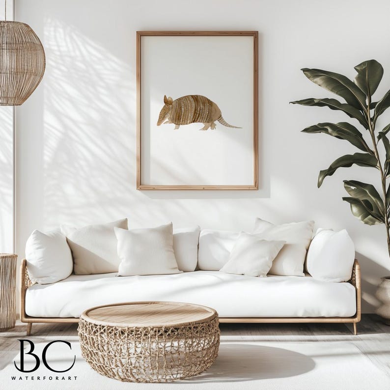 May include: A watercolor illustration of an armadillo in shades of brown, framed in a light wood frame and hanging on a white wall above a white couch with beige throw pillows. A woven wicker coffee table sits in front of the couch.