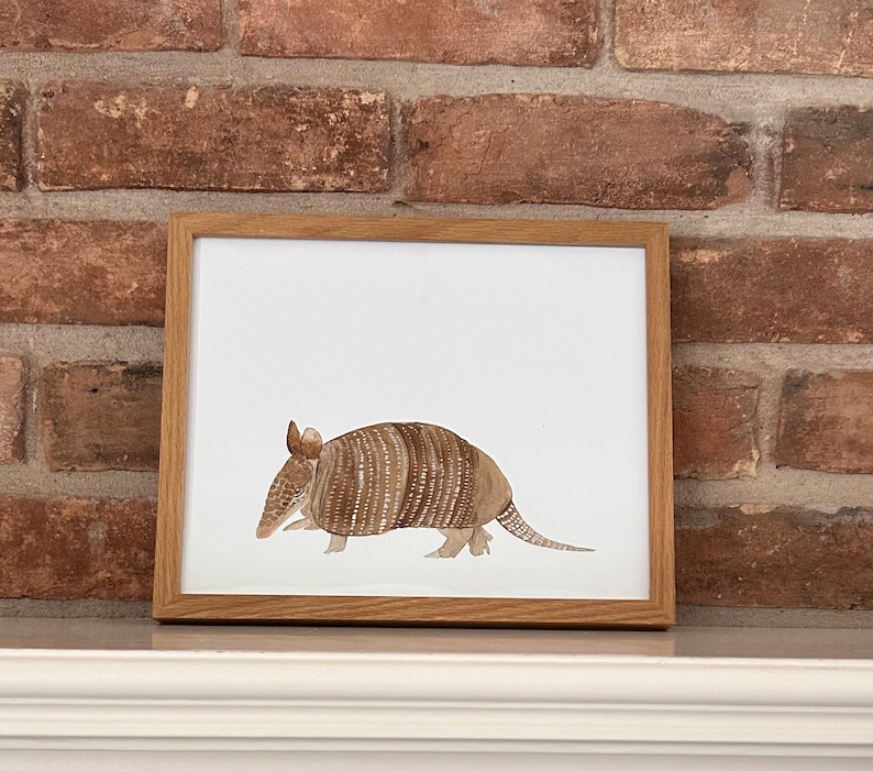 May include: A framed watercolor painting of a brown and tan armadillo on a white background. The armadillo is walking to the right and has a long tail.