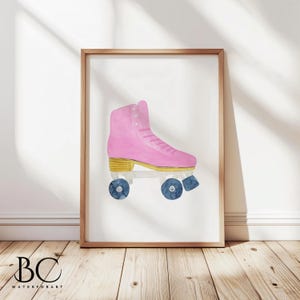 May include: A framed watercolor print of a pink roller skate with gold accents and blue wheels. The artwork is displayed on a white background and framed in a light brown wooden frame. The print is displayed on a wooden floor.