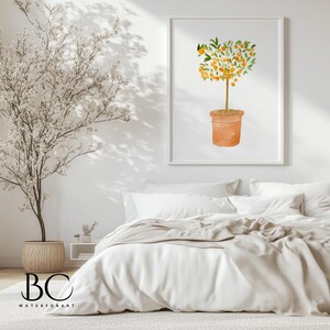 May include: A watercolor painting of a small orange tree with a terracotta pot. The tree is in a white frame and is hanging on a white wall above a white bed with a beige throw blanket.