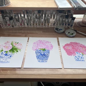 Puede incluir: Tres pinturas de acuarela de flores rosas en jarrones azules y blancos. Los jarrones tienen patrones intrincados. Las pinturas est&aacute;n sobre papel blanco, y el fondo incluye materiales de arte como tubos de pintura y frascos.