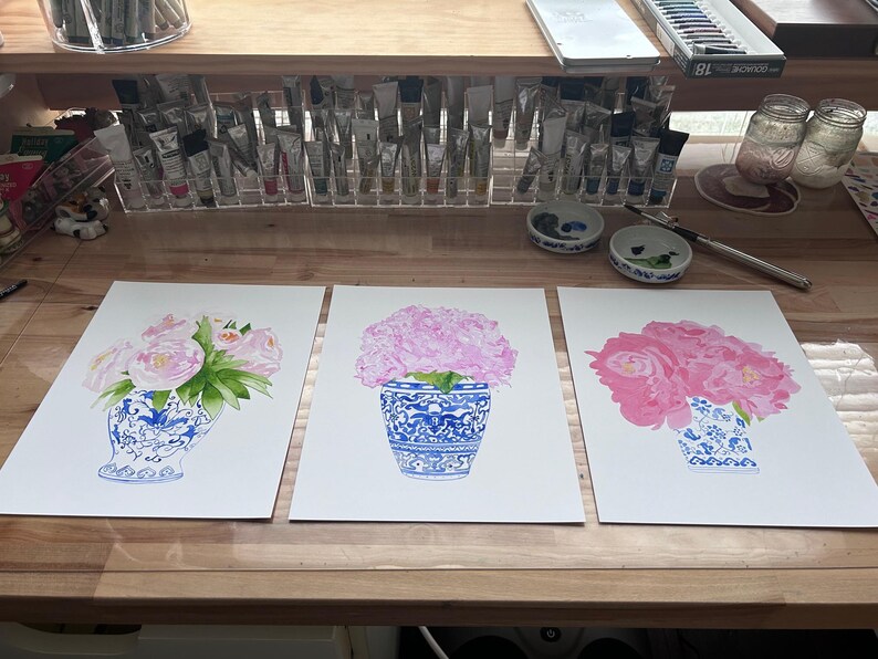 Puede incluir: Tres pinturas de acuarela de flores rosas en jarrones azules y blancos. Los jarrones tienen patrones intrincados. Las pinturas est&aacute;n sobre papel blanco, y el fondo incluye materiales de arte como tubos de pintura y frascos.