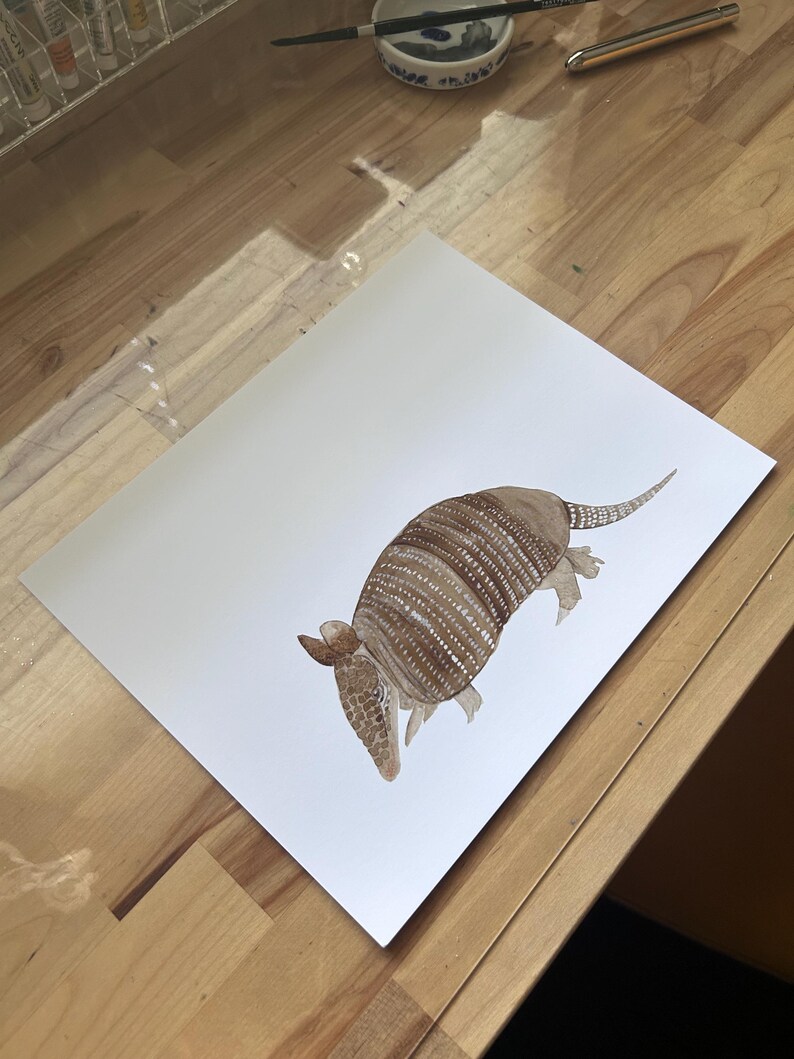 May include: A watercolor painting of an armadillo on white paper. The armadillo is brown and has a detailed shell with white stripes. The painting is on a wooden surface.