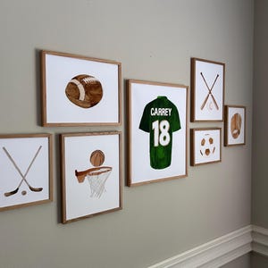 May include: A collection of framed watercolor art prints featuring sports themes. Includes a football, basketball, hockey sticks and puck, baseball bat and ball, baseball glove, soccer ball, and a green jersey with the name "Carrey" and the number "18".