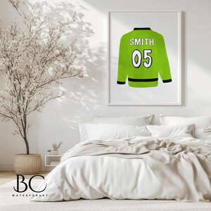 Puede incluir: Una camiseta de hockey verde y negra con el nombre "SMITH" y el n&uacute;mero "05" en la espalda. La camiseta est&aacute; colgada en una pared blanca en un dormitorio.