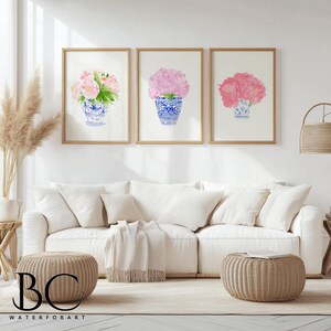 Puede incluir: Tres impresiones enmarcadas en acuarela de flores rosas en jarrones azules y blancos. La obra de arte se exhibe sobre un sof&aacute; blanco con cojines y una otomana tejida. El tema general es floral y decoraci&oacute;n del hogar.