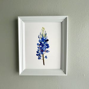 May include: A white framed print of a watercolor painting of a bluebonnet flower. The flower is blue with a yellow center and is set against a white background.