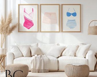 May include: Three watercolor illustrations of swimwear on a white background. The illustrations are framed in natural wood frames. The illustrations include a pink swimsuit, blue and white striped swim bottoms, and a blue bikini top.