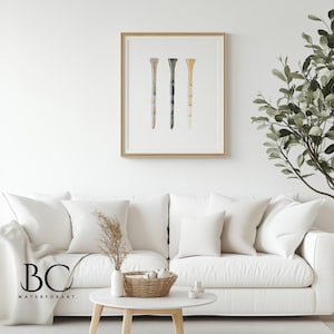 May include: A watercolor painting of three golf tees in different colors: beige, gray, and brown. The tees are arranged vertically in a row. The painting is framed in a light wood frame.