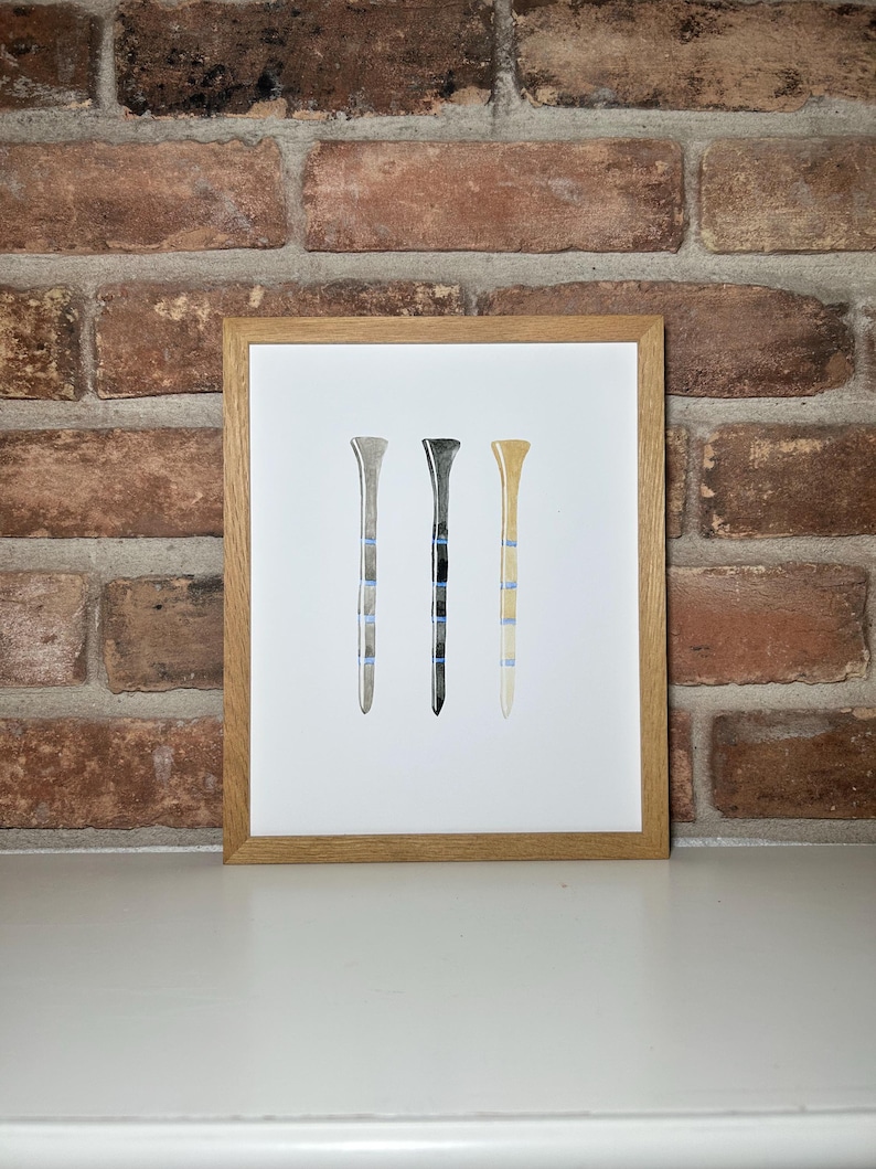 May include: A framed print of three watercolor golf tees in different colors: gray, black, and beige. Each tee has blue stripes.