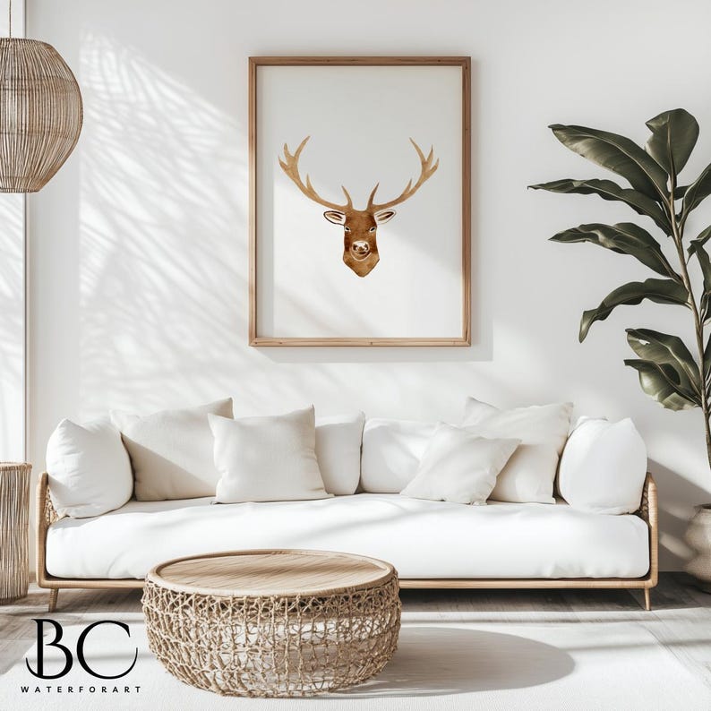 May include: A watercolor painting of a deer head with antlers in a light brown frame hanging on a white wall above a white couch with white pillows. A woven wicker coffee table sits in front of the couch.